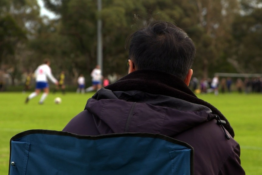 The back of a man sitting on a folding chair on the sideline of a soccer game. Players can be seen in the distance.